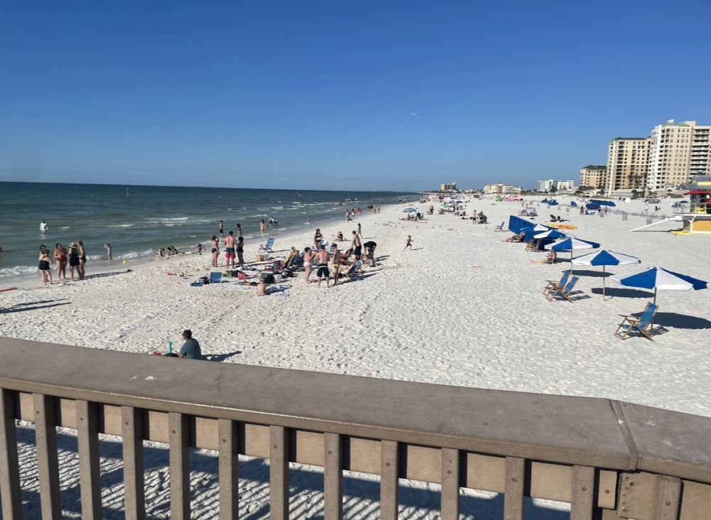 View from a walkway over the beach: rail across the foreground, the white-sand beach stretching out as far as the horizon, a scattering of people and umbrellas and beach chairs. A few tall appartment buildings partly visible on the right, the sea on the left.