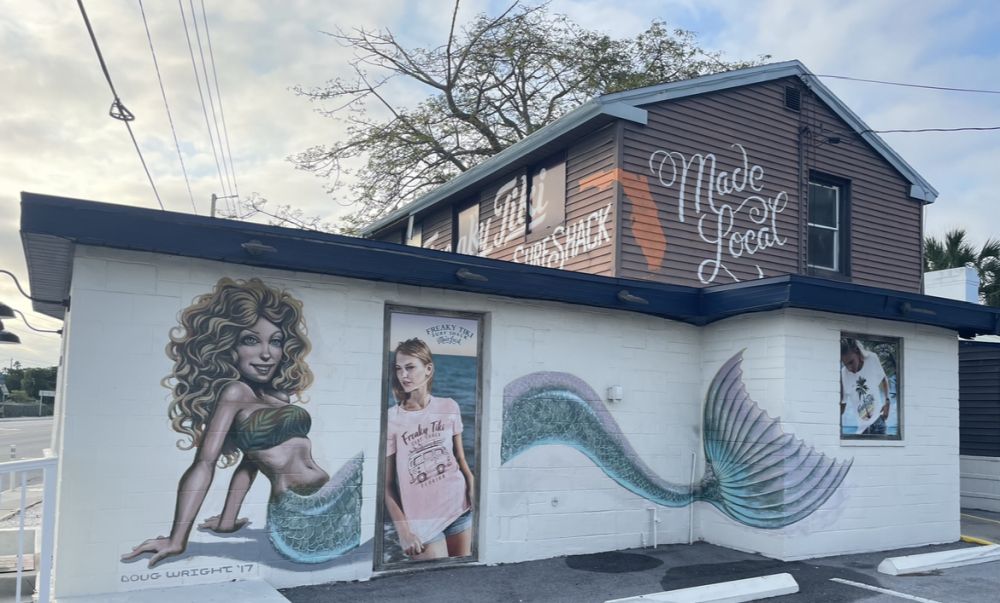 Side of a building with an image of a mermaid and also a woman in a t-shirt leaning in a doorway. Her t-shirt and the sign on the building read "Freaky Tiki Surf Shack"