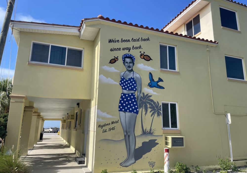 Side of a building painted light yellow, with a mural depicting a 1940s woman in a bathing suit and the words "We've been laid back since way back" and "Keystone Motel Est. 1945."