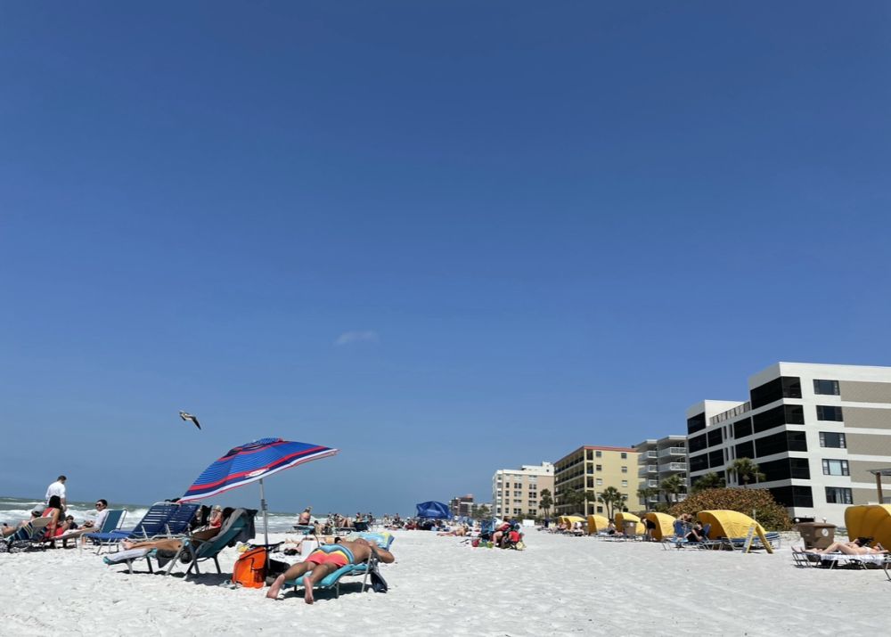 View down a white-sand beach, buildings on the right, people lying on beach chairs on scattered across the beach.