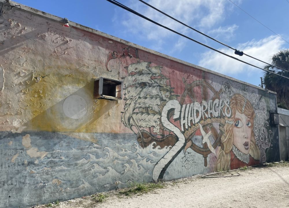 Side of a building painted with a mural that shows the face of a woman and also a sailing ship, with the word Shadracks.