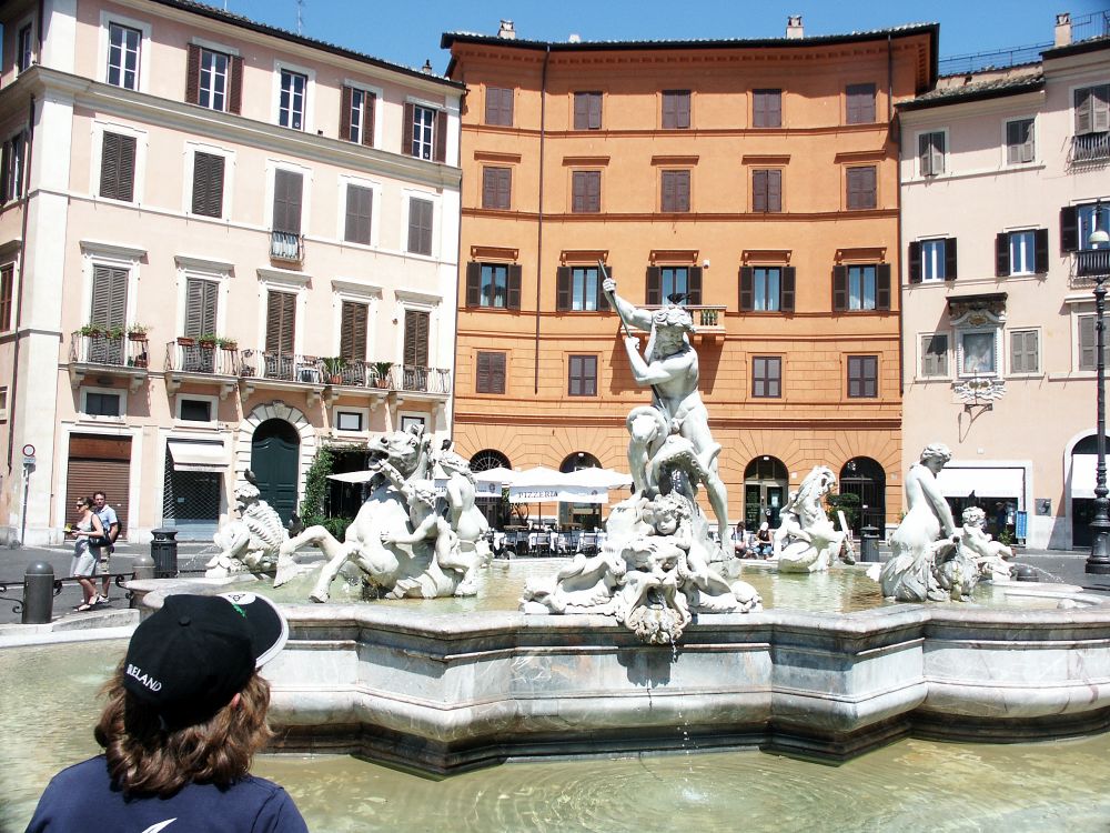 A fountain with three buildings behind it. The fountain is very ornate with an image of Neptune in the middle and various sea creatures and horses rising out of the water.