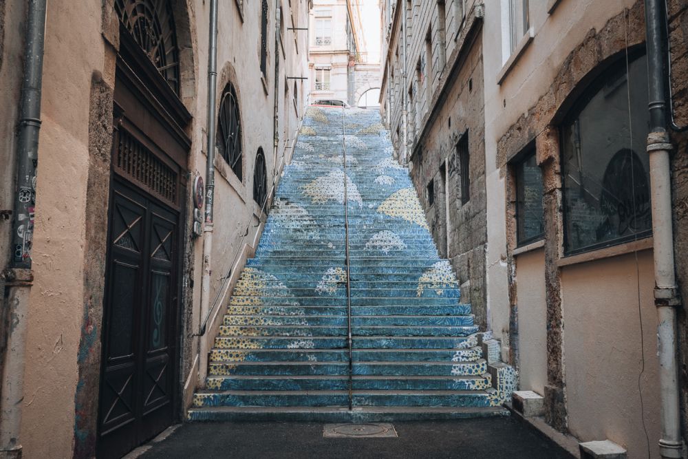 A view up a staircase between two tall buildings. The staircase is painted in a subtle pattern.