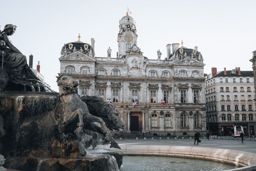 A baroque fountain in the foreground with horses emerging from the stone. A very ornate palace-like building in the background across the plaza.