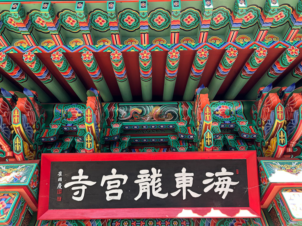 Looking up under the eaves of a temple, wooden beams fancifully decorated with painted flowers, a dragon and other figures.