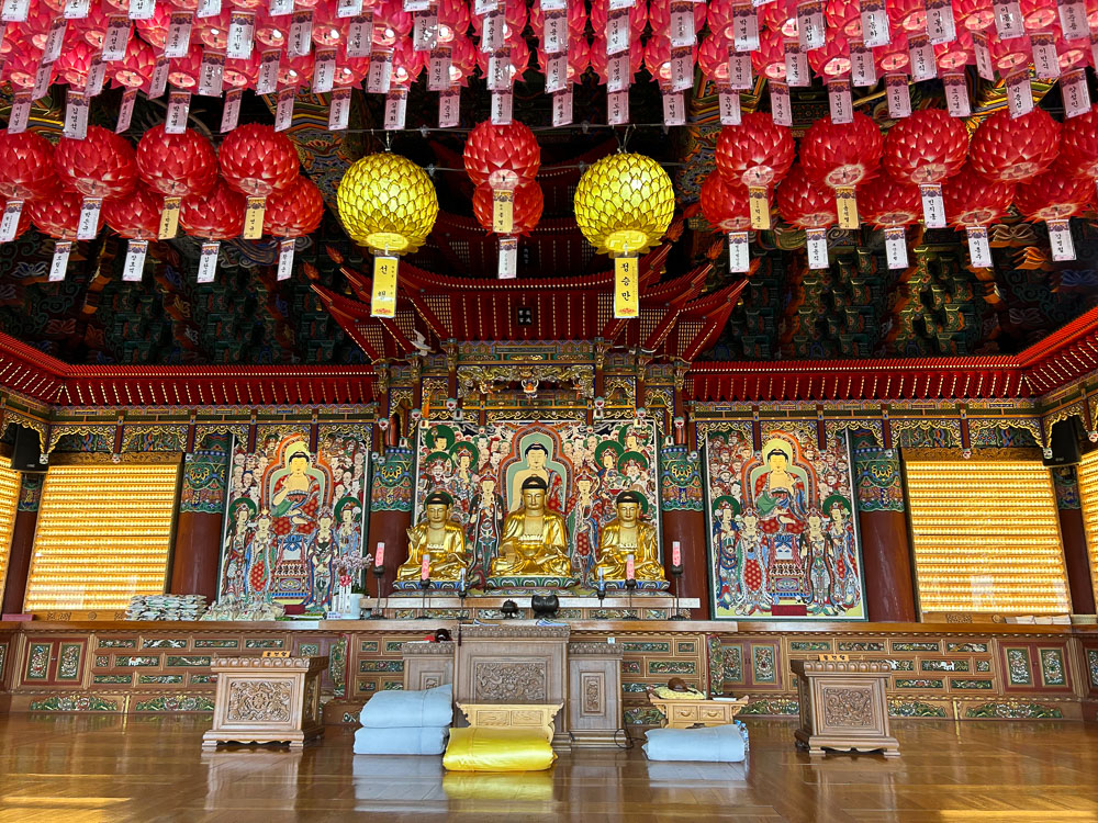 A shrine with three central golden statues of the Buddha and colorful painted images behind and beside them. red paper lanterns hang from the ceiling.