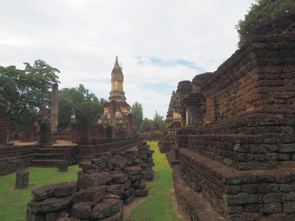 A row of stupas in dark red laterite stone on the right. In the middle, a low ruined wall. A platform on the right with the remains of columns, and a large lotus-top stupa behind.