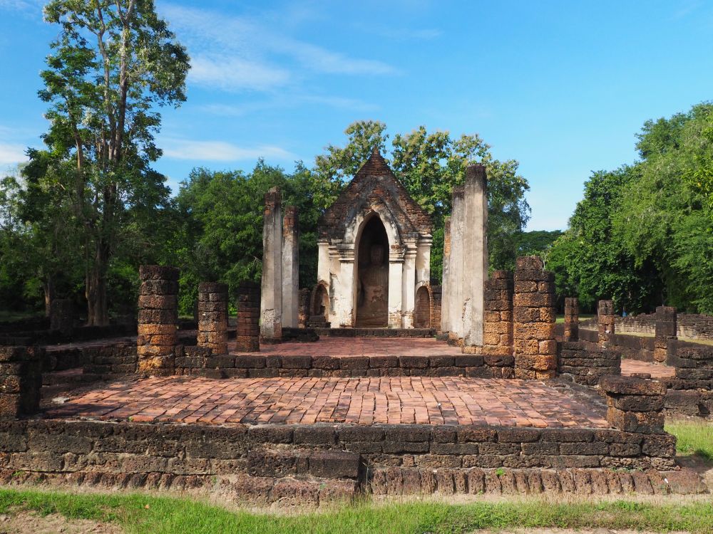 A very simple temple, like a house with a peaked roof and an arched doorway. In front of it, a paved floor area with the remains of columns on either side.