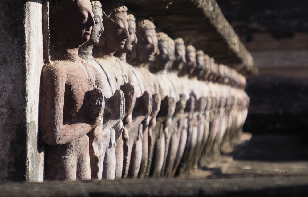 Looking down a row of carvings of monks, each holding hands together in prayer in front of them. The nearest is in focus, while the rest are blurry.