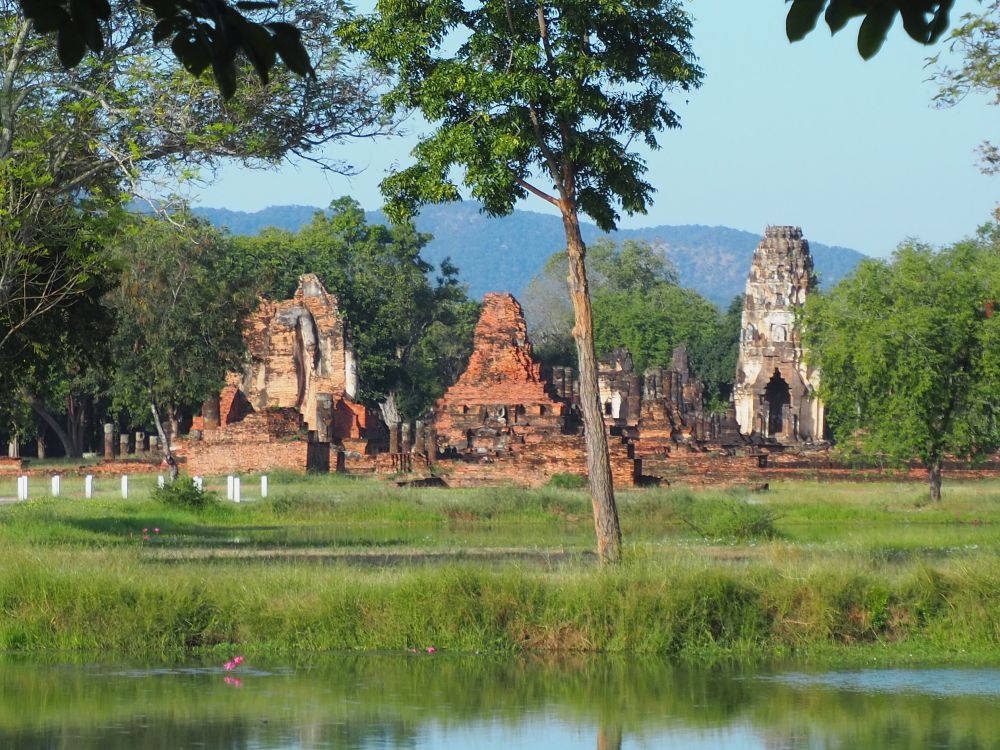 Seen across a moat and a grassy field, 3 buildings in varying states of decay. On the right is a prang, in the middle a stupa that's reduced to brick, and on the right a somewhat more intact stupa.