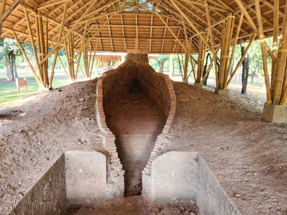 Under a wooden roof open at the sides, what remains is a mound with an oblong opening in it lined with bricks and the base of what was once a chimney at the far end.