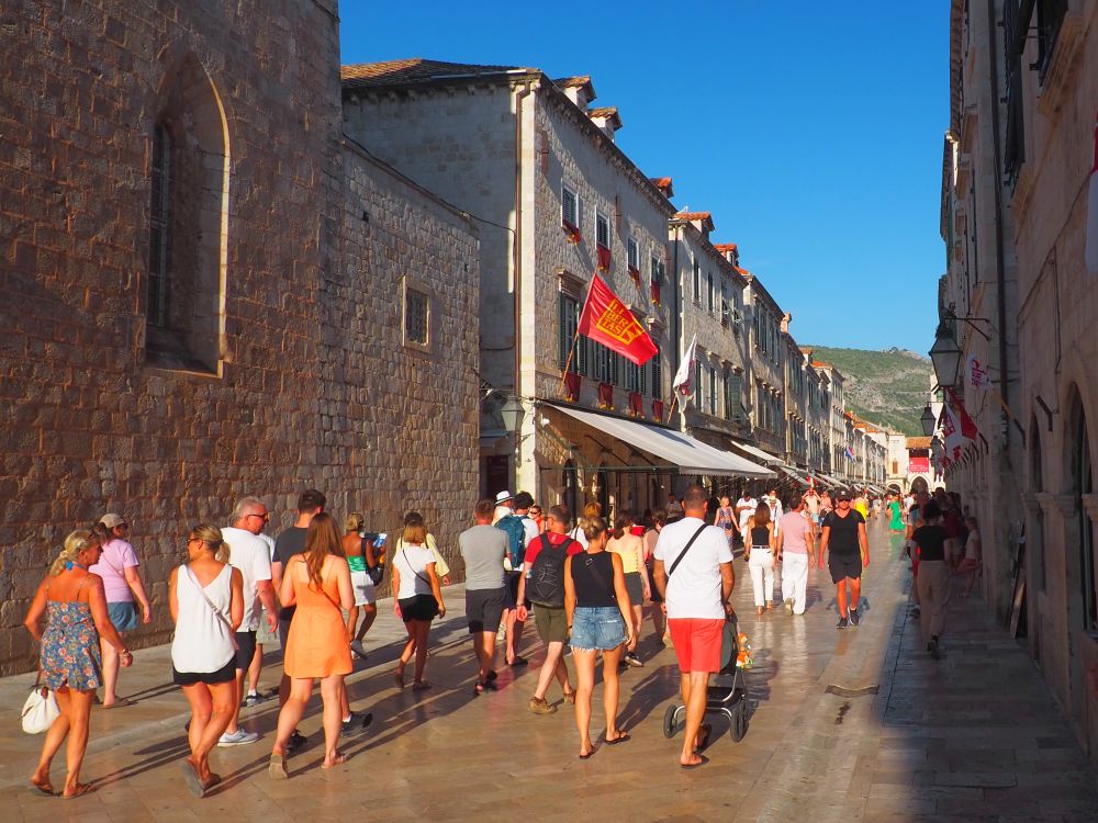 A wide street paved with flat stones, shops on either side, lots of people walking along it.