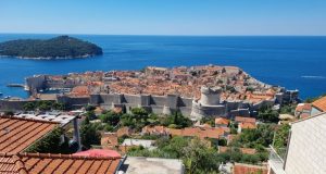 A view of Dubrovnik from a hill: red-roofed buildings, a wall around it with a fort on the near side, the blue sea beyond the city.