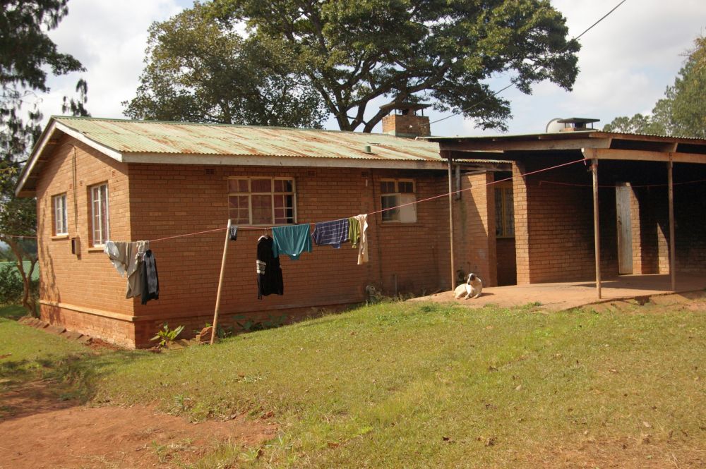 A single-story brick house with a corrugated-iron roof. In front is a small place with a roof over it and laundry hanging in the sun next to it.