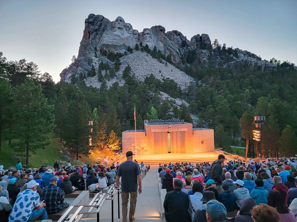 An outdoor amphitheater with lots of people sitting, backs to the camera, facing the darkening Mount Rushmore. A small building on the stage is lit up.