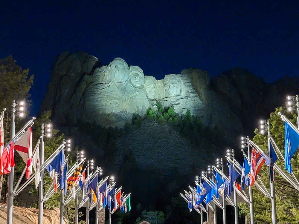 View of the image of 4 presidents, lit up at night. The view sights down the Avenue of Flags, so the brightly colored flags lining the path seem to point to the image.