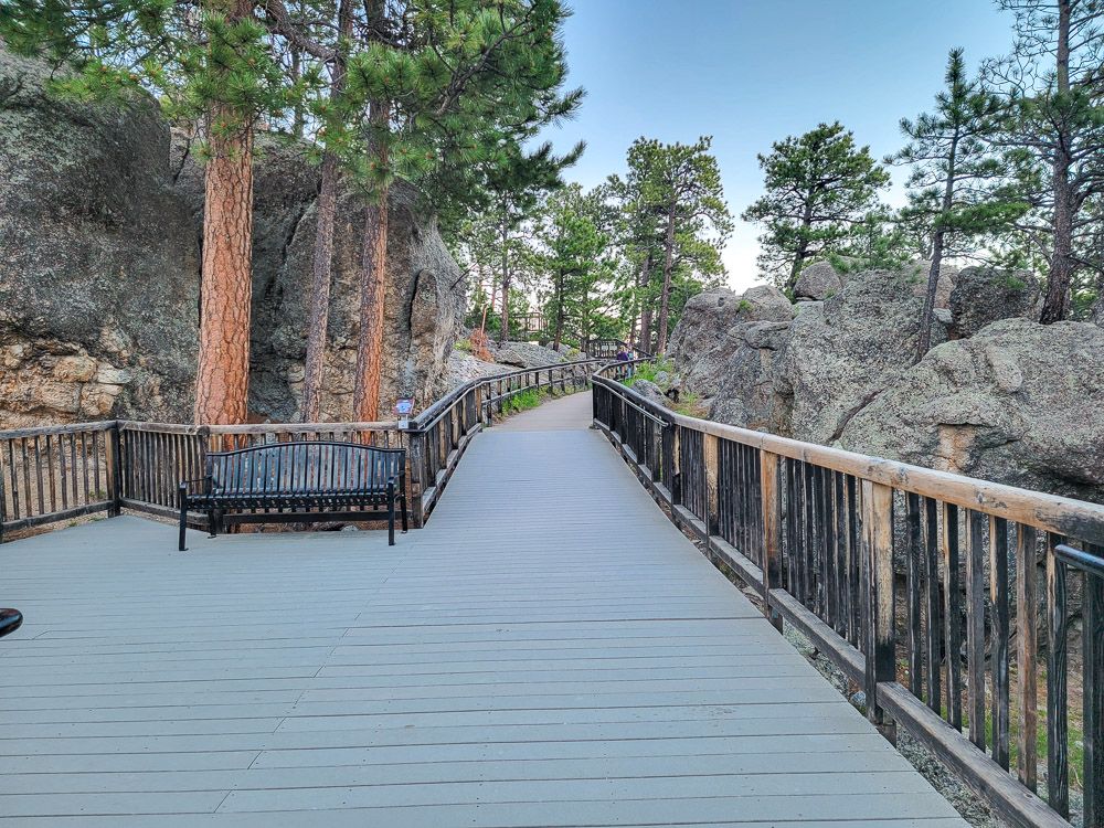 The trail is a wide wooden boardwalk - at least in the section shown - with railings along both sides and a park bench. On either side are large rocks and pine trees.