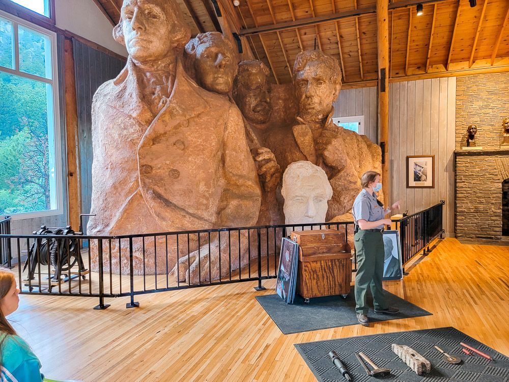 A park ranger stands in front of a small (ceiling high) version of the Mount Rushmore image of the Presidents. She is speaking to an off-camera audience.