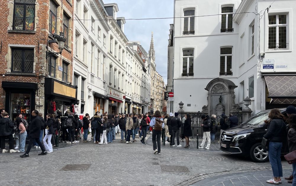 Looking down a street with shops and apartments - a crowd of people milling around. On the corner, the fenced-off Manneken Pis fountain.