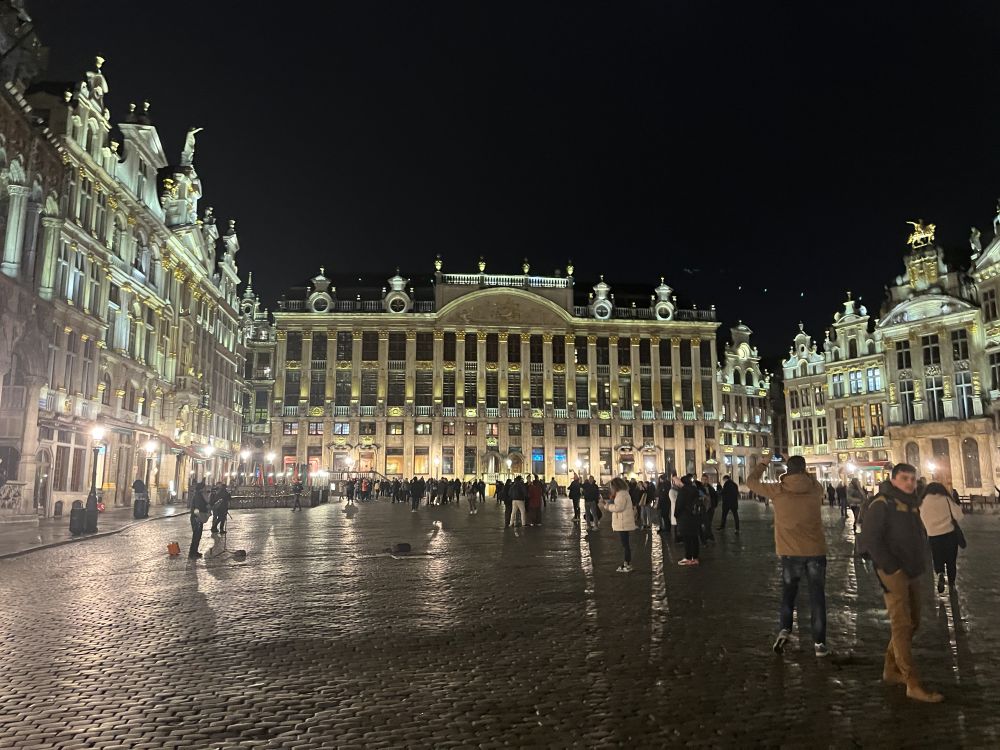 A large plaza at night with ornate buildings around the 3 visible sides, lit up so that the gold-colored details shine. 