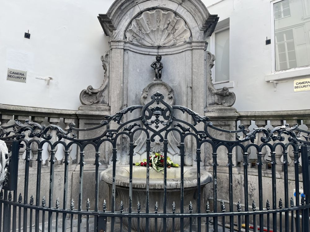 The Manneken Pis stature sits in the center of a rather baroque stone fountain. It is in the corner of two buildings and a metal fence surrounds it.