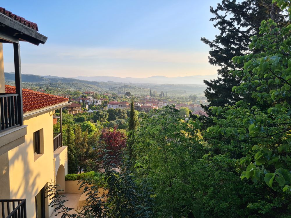 Between the side of the hotel and some trees, a huge view of a green valley below, with a mountain range in the distance beyond that.