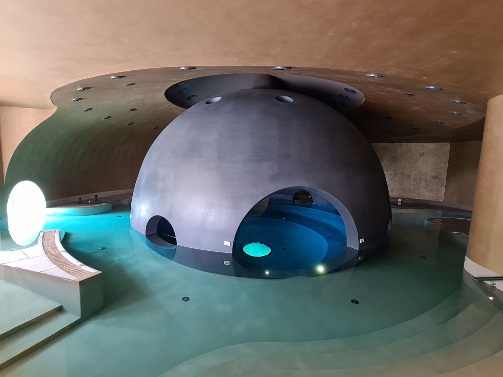 Blue water, lights shining from underneath, a low ceiling and, in the center, a large grey spherical shape with circular openings in its side.