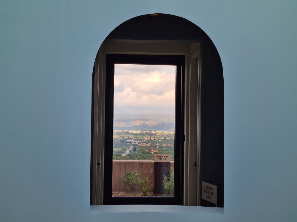 An arched window on a gray wall opens into a hallway where a door stands open with a view over the valley below.