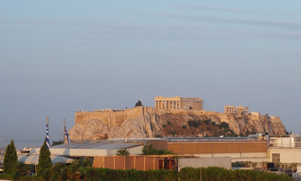 The acropolis sitting on a rocky hill with the tops of lots of buildings visible between the camera and the hill.