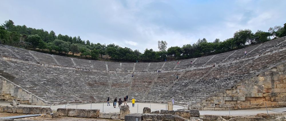 A half-circle of seating built against a hill, with trees around the top, and some people at the bottom on what would have been the stage.