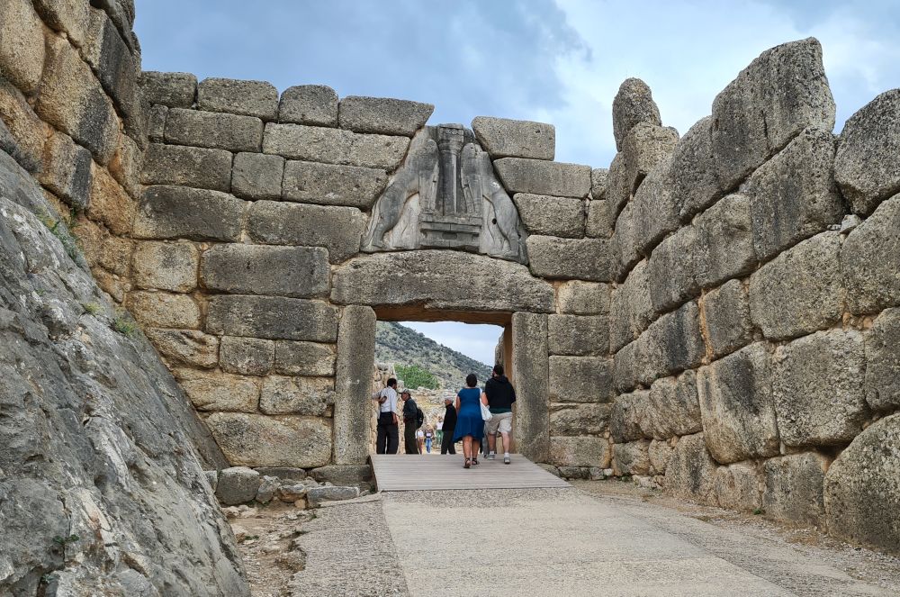 Large stone walls frame a road leading through a large square gateway with people inside the gateway (the height of the gateway is about twice the height of the people.