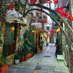 A narrow street with buildings along both sides and lots of potted plants and flowers in front of them, as well as a bougainvillea over the street.