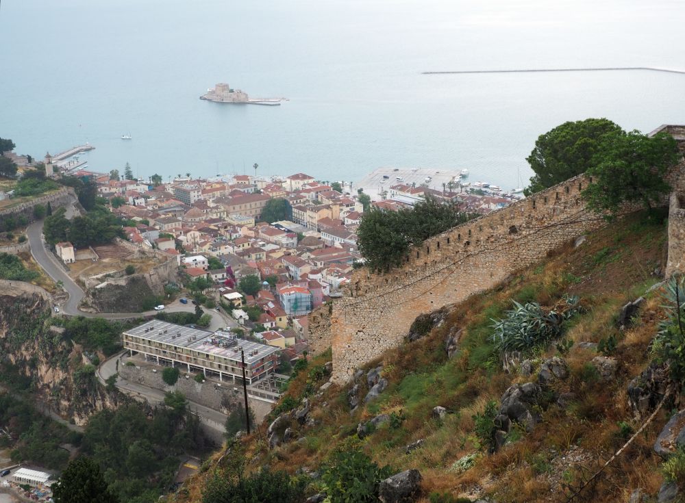 A wall of the fortress in the near distance; beyond that, far below, the cluster of buildings that is old Napflio, and, on the blue water beyond that, a small island filled by a small fortress.