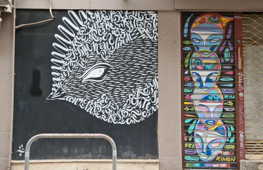 Street art: white on black, a figure of a fish composed of lots of symbols and letters. Beside it, on a shop's shutter, another image, more colorful, of a series of abstract faces. 