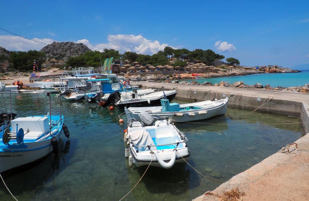 A row of small fishing boats along a concrete pier. Beyond them, the island, which is rocky, with rows of thatched sun shelters along its rocky edge, and trees behind them.