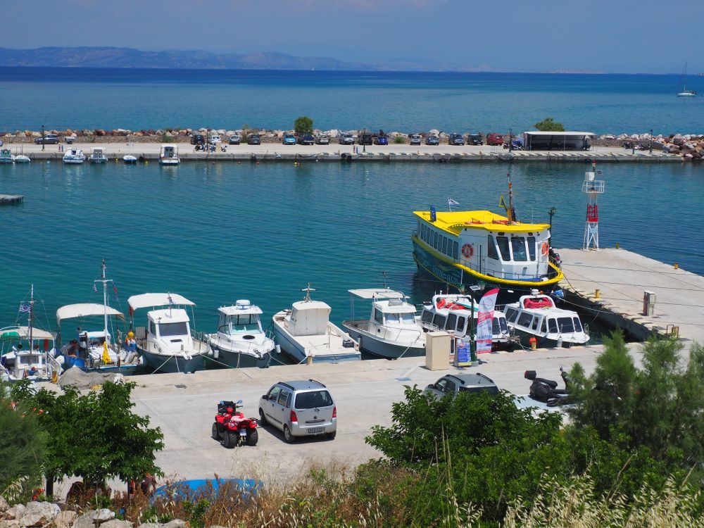 Looking down on a small harbor with a line of small boats along a concrete pier and, on a larger pier, a larger boat with a yellow roof.