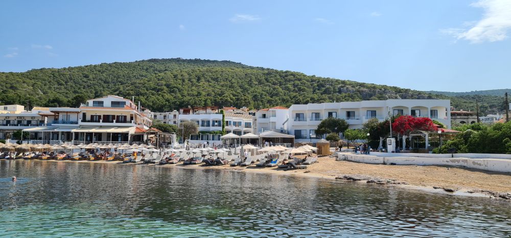 A cluster of buildings seen from across very calm water - all are low-rise, and the beach in front of them is mostly covered with lounge chairs and umbrellas.