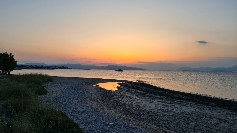 Sand and pebble beach in the foreground, the sunset, with a boat in front of it, in the background.