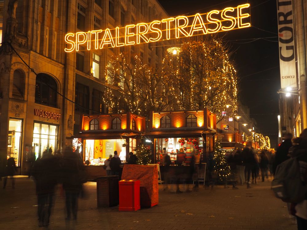 A large sign stretched across the street in white lights reads "Spitalerstrasse." Beyond that, a city street with lit-up shops on either side and a row of market stalls down the middle.