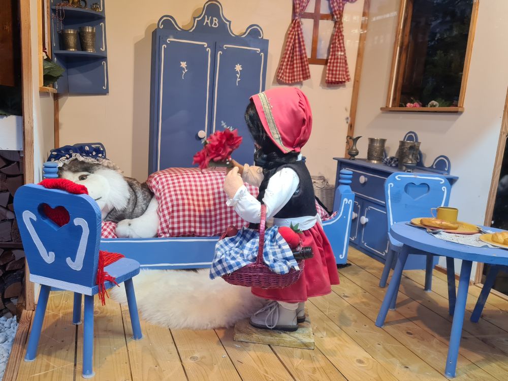 Doll of Little Red Riding Hood in the center, holding a red bouquet. Blue-painted furniture, including the bed that the wolf lies on, under the covers.