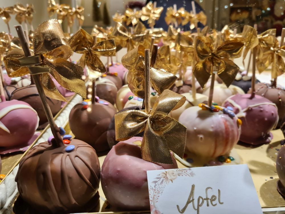 A table filled with rows of apples that have been dipped in, presumably, chocolate, mostly in pinks, whites or browns. Each stick has a gold bow tied around it.