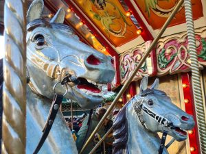 Two painted white horses - just their heads - on a merry-go-round with the colorful painting of the center part of the carousel visible behind them.