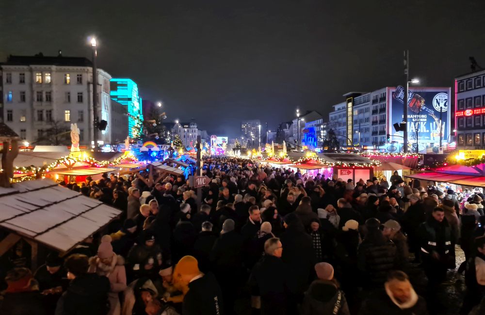 A huge crowd of people down the length of the rectangular space, with brightly lighted stalls along the sides and neon on the buildings on either side.