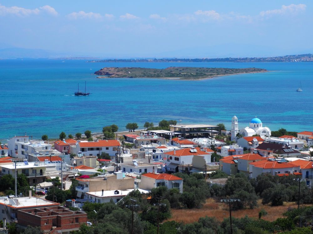 A view of the small town of Skala, all low buildings of just a few stories. The blue dome of the church stands out. Beyond the town, the sea a small flat island.