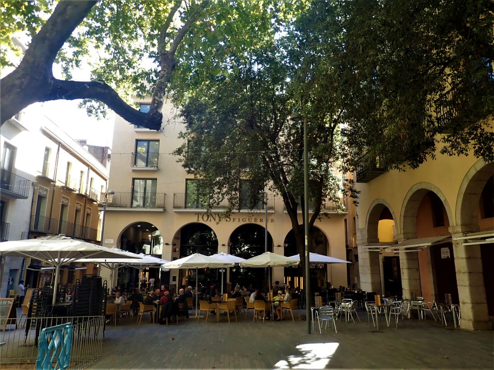 A plaza with cafe tables set out with umbrellas over them and trees shading them too - Tony's Bar is visible behind the tables.