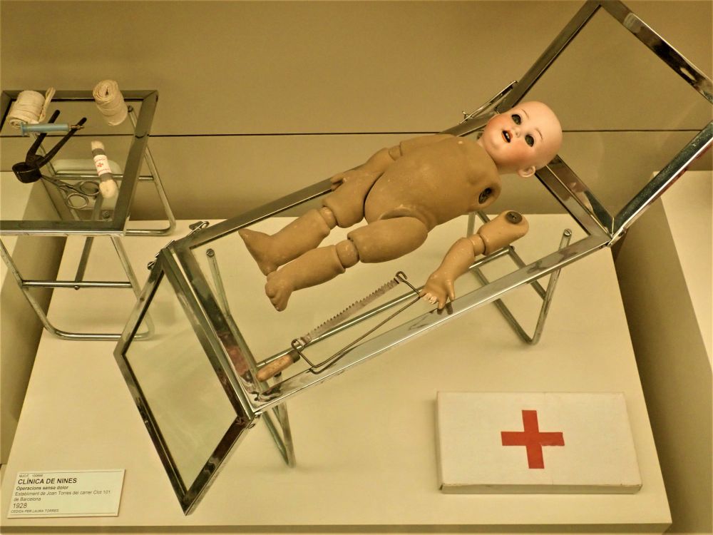 A naked doll with no hair lies on a sort of stretcher, its arm lying next to it. Various medical equipment is arranged around the stretcher.