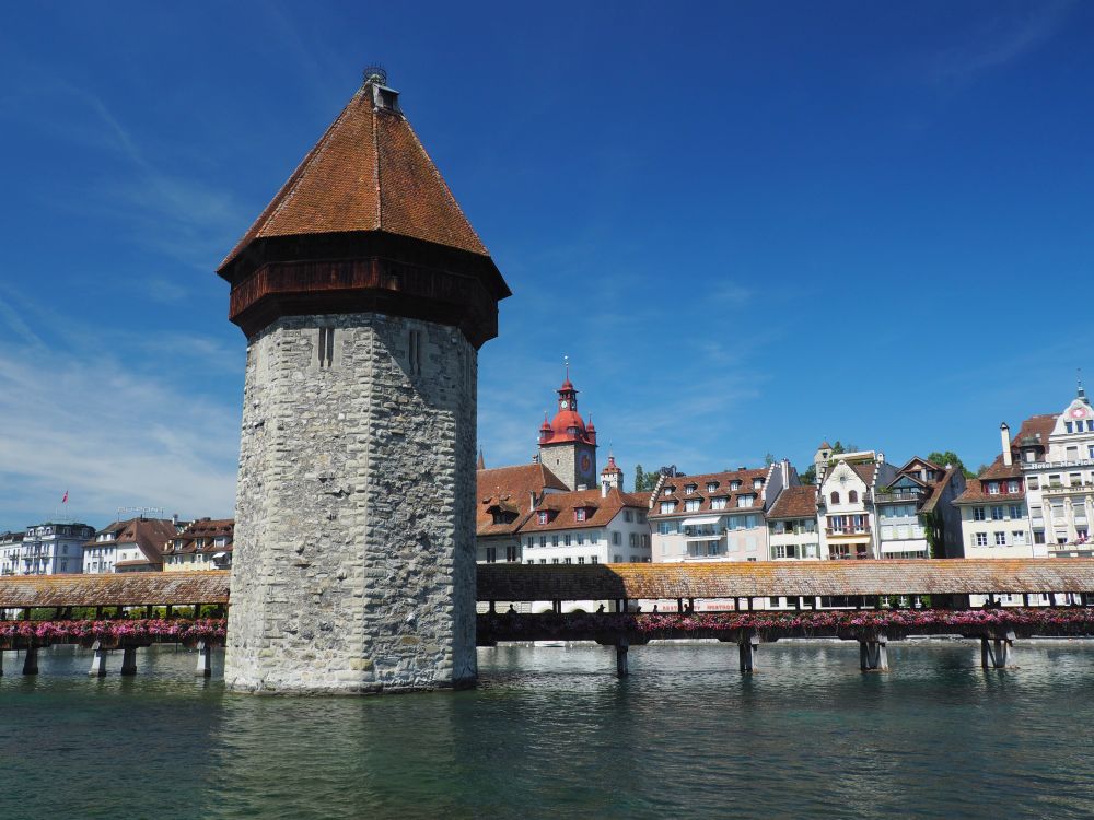 A covered bridge crossing a river, with a large stone tower in the middle, topped with a pointy cap roof.