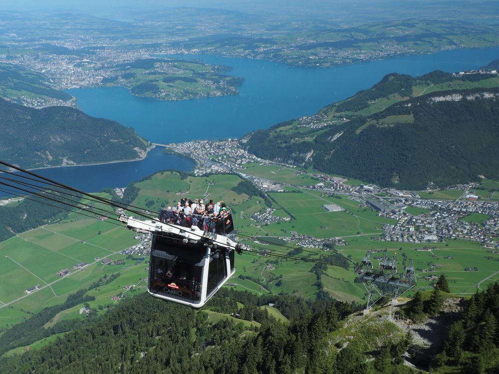 A cable car in midair, a group of people visible on its roof. Behind, a far view of Lake Lucerne.