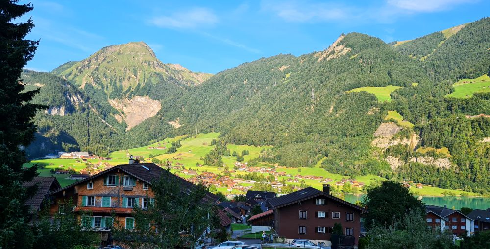 In the near distance, several chalet-type houses. Beyond them a green valley with more scattered houses, and mountains in the background.