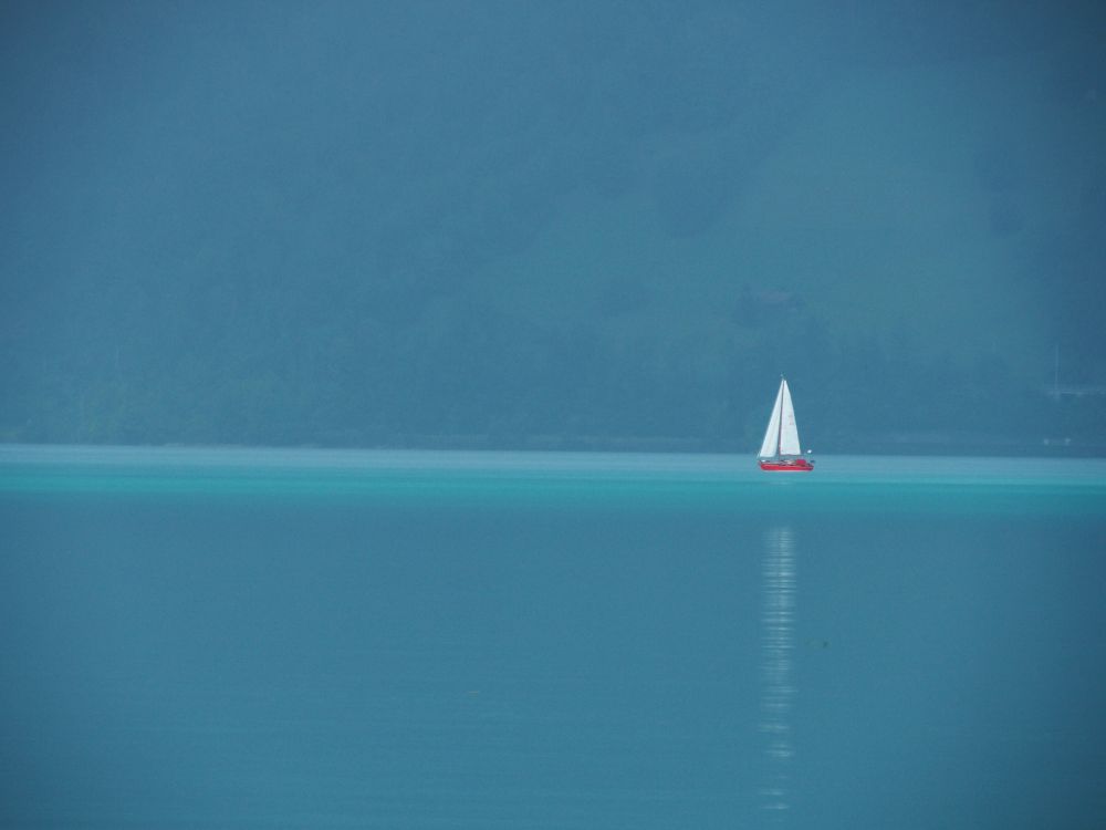 Blue water, blue background (mountains in the mist) and a single red sailboat on very still water.